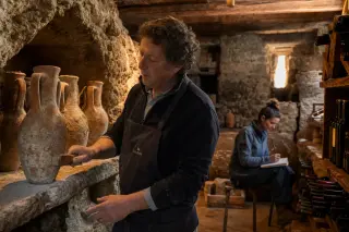 Wine cellar combining Roman and Alpine elements, winegrower brushing amphorae, young woman sketching vine motifs in warm Mediterranean afternoon light.