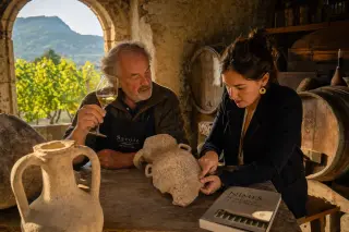 Elderly winemaker and young historian study ancient amphorae together in a sunlit stone wine cellar, with wine barrels, alpine vines, and hints of Roman architecture in a warm, candid atmosphere.