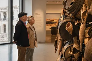 Two elderly locals in a modern museum gallery appreciate an installation combining Roman amphorae fragments and Savoyard wine barrels, bathed in warm Mediterranean light.