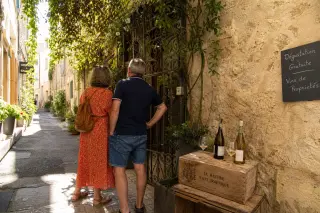 Curious locals explore a vine-covered gate in a sunlit Nîmes alley, with wine glasses nearby and warm Mediterranean light.