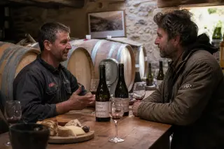 Two winemakers in discussion at a rustic cellar counter with wine glasses, cheeses and soft sunlight