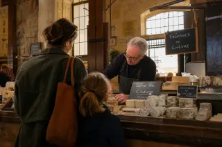 Artisan cheesemonger arranging products behind a market counter as a mother and daughter observe, in warm morning light inside Halles de Chambéry.