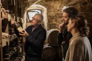 Vintner selecting a bottle in a dimly lit stone cellar as visitors observe, soft golden light illuminating old bottles and barrels.