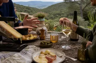 Unfinished raclette meal with garrigue honey, Alpine wine, and two friends' hands sharing food on a rustic terrace with sunlit Mediterranean hills in the background.