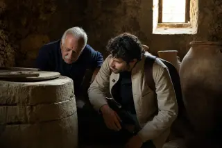 Elderly winemaker and young visitor exploring textures on ancient barrel in softly lit historic wine cellar, with warm sun patches highlighting stone walls and amphorae.